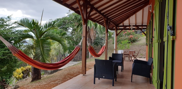 Een overdekt terras met rieten meubilair, een hangmat en uitzicht op tropische begroeiing.