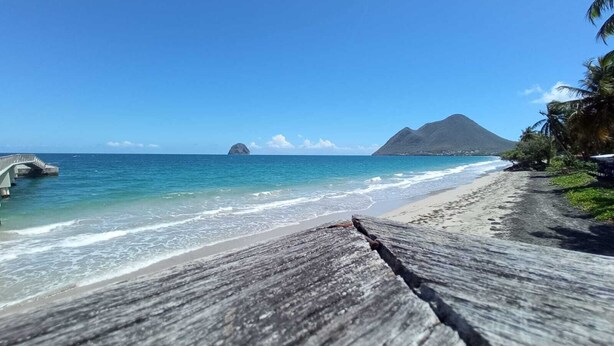 Une plage avec une promenade en bois, une eau d’un bleu limpide et une montagne en arrière-plan.