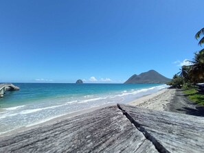 Une plage avec une promenade en bois, une eau d’un bleu limpide et une montagne en arrière-plan.