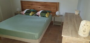 Quarto com cabeceira de madeira, colcha verde, duas mesas de cabeceira com luminárias e uma mesinha de cabeceira com objeto decorativo.