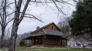 A rustic log cabin with a porch, surrounded by trees and a snowy landscape. - JuneBug Log Cabin, Nashville Music History, Dreamy Loft Suite, Stone Frpl. (Hendersonville)