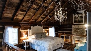 A rustic bedroom with a large bed, a chandelier, a fireplace, and a stone wall. - JuneBug Log Cabin, Nashville Music History, Dreamy Loft Suite, Stone Frpl. (Hendersonville)