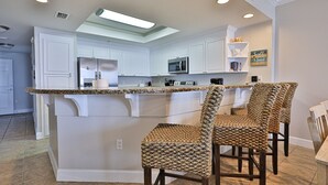A kitchen with white cabinets, a granite countertop, and a built-in microwave. There are two woven chairs at the island.