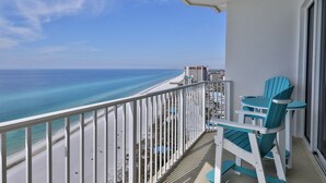 A balcony with a view of the ocean and beach, featuring a white railing and two turquoise chairs.