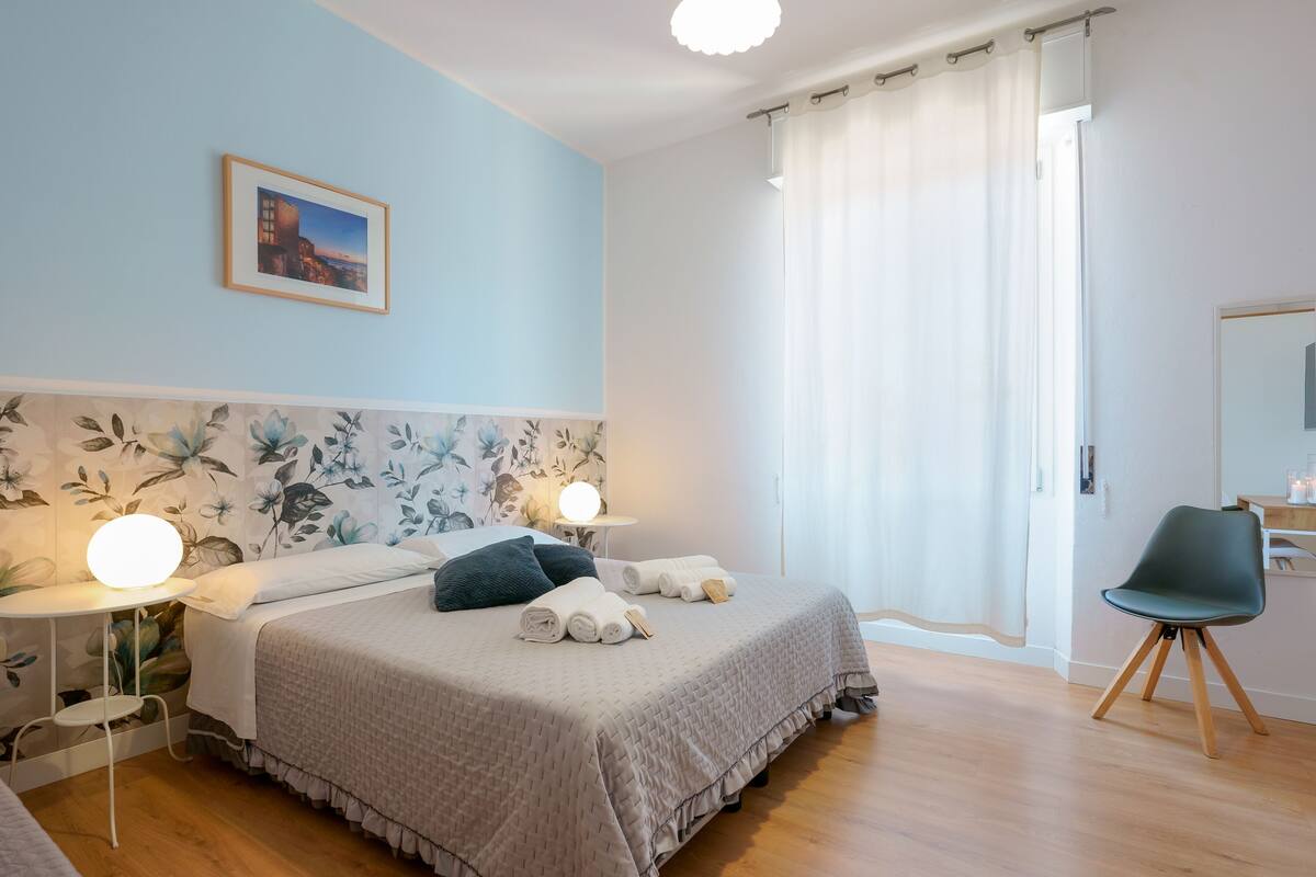 Une chambre à coucher comprenant un lit, des tables de chevet, une chaise, une lampe fixée au mur et une fenêtre avec des rideaux.