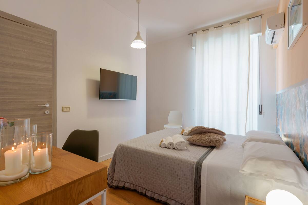 Une chambre d’hôtel avec un lit, un téléviseur fixé au mur, un bureau avec des bougies et une fenêtre avec des rideaux.