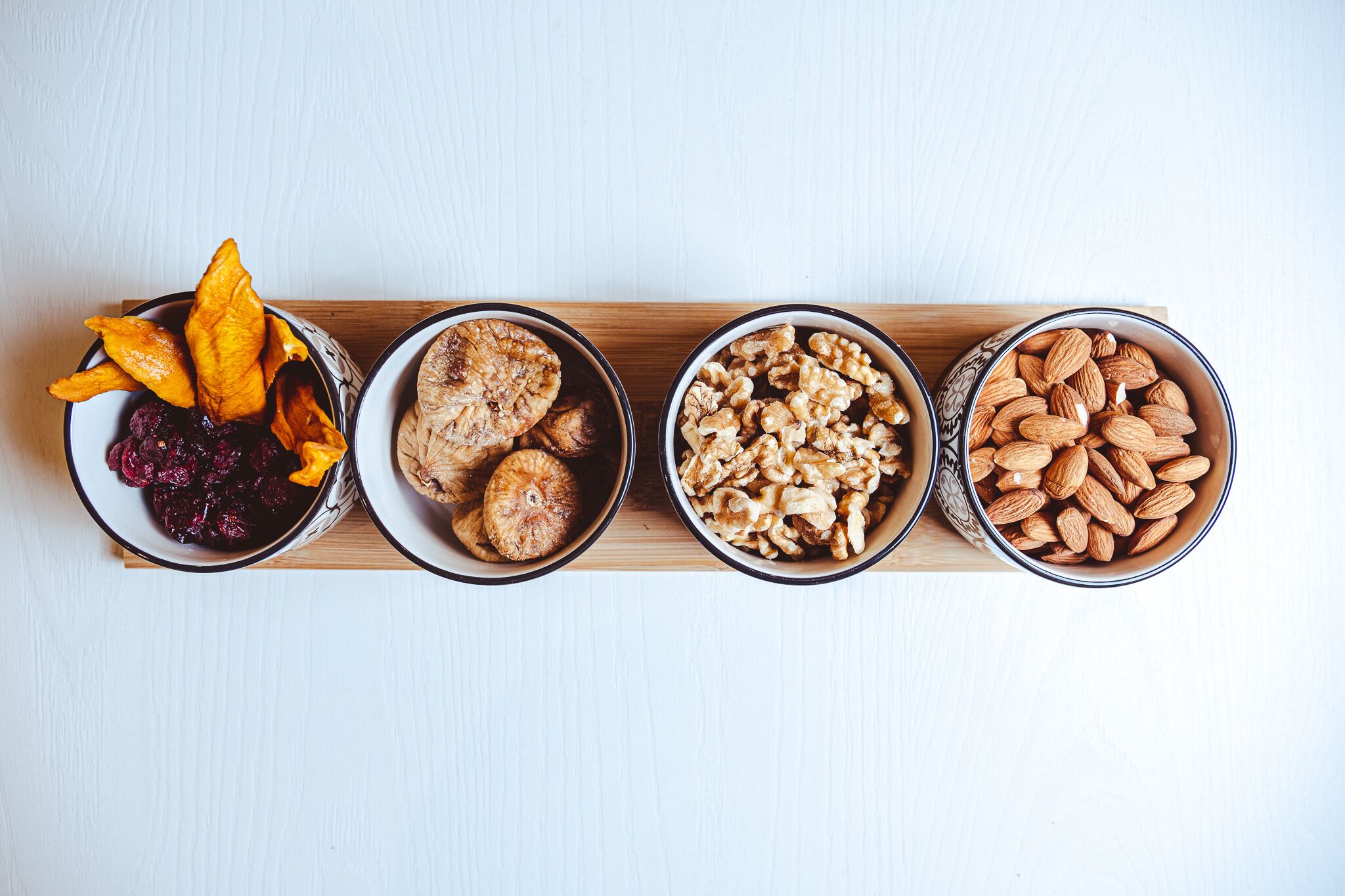 Cuatro recipientes con frutos secos y frutas deshidratadas sobre una tabla de madera.