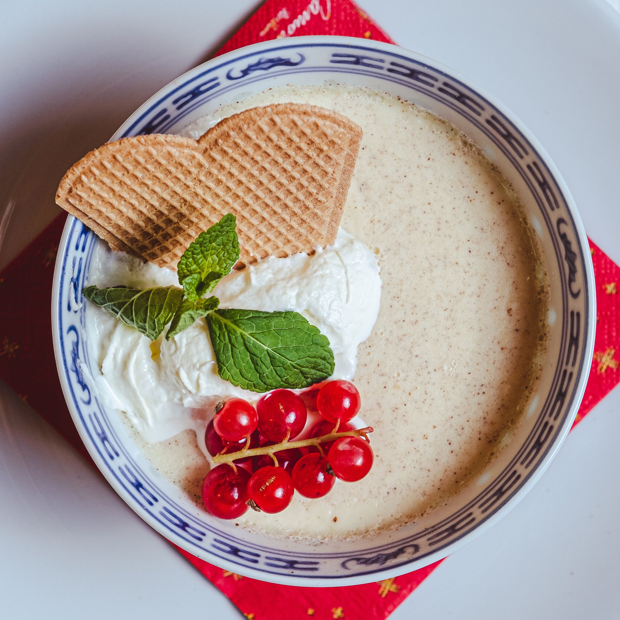 A plate with a creamy dessert, a slice of wafer, whipped cream, mint leaves, and red currants.