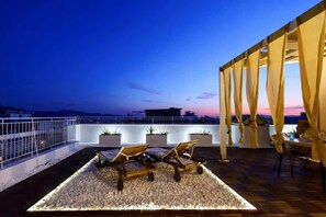 Una terraza en la azotea con dos sillones de descanso, una zona de comedor y vistas al horizonte de la ciudad al atardecer.