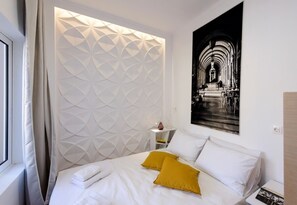 Een moderne hotelkamer met een hoofdbord in geometrisch patroon, een bed met witte lakens en mosterdgele kussens, een klein nachtkastje met een vaas, en een ingelijst fotoportret aan de muur.