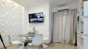 A modern living room with a flat-screen TV mounted on the wall, a dining area with white chairs and a glass table, and a white bookshelf.
