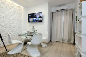 A modern living room with a flat-screen TV mounted on the wall, a dining area with white chairs and a glass table, and a white bookshelf. - City Skyloft - Top location & Private Roof Garden (Kalamata)