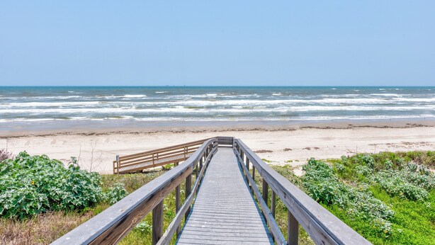 Ein hölzerner Steg führt zu einem Strand, im Hintergrund sind Wellen zu sehen.