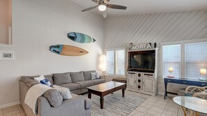Ein Wohnzimmer mit einem grauen Ecksofa, einem Holztisch und einer TV-Konsole mit der Aufschrift 'AORTA'.