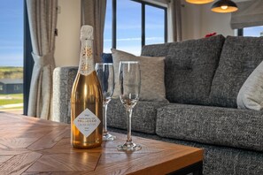 A bottle of Prosecco on a wooden table with two glasses beside it, in a room with a grey sofa and a view of the outdoors. - Hiraeth - Luxury Lodge With Hot Tub Close to Beach (Tenby)