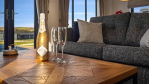 Ein Holztisch mit einer Champagnerflasche und zwei Gläsern, ein graues Sofa mit Kissen und ein Blick ins Freie durch große Fenster.