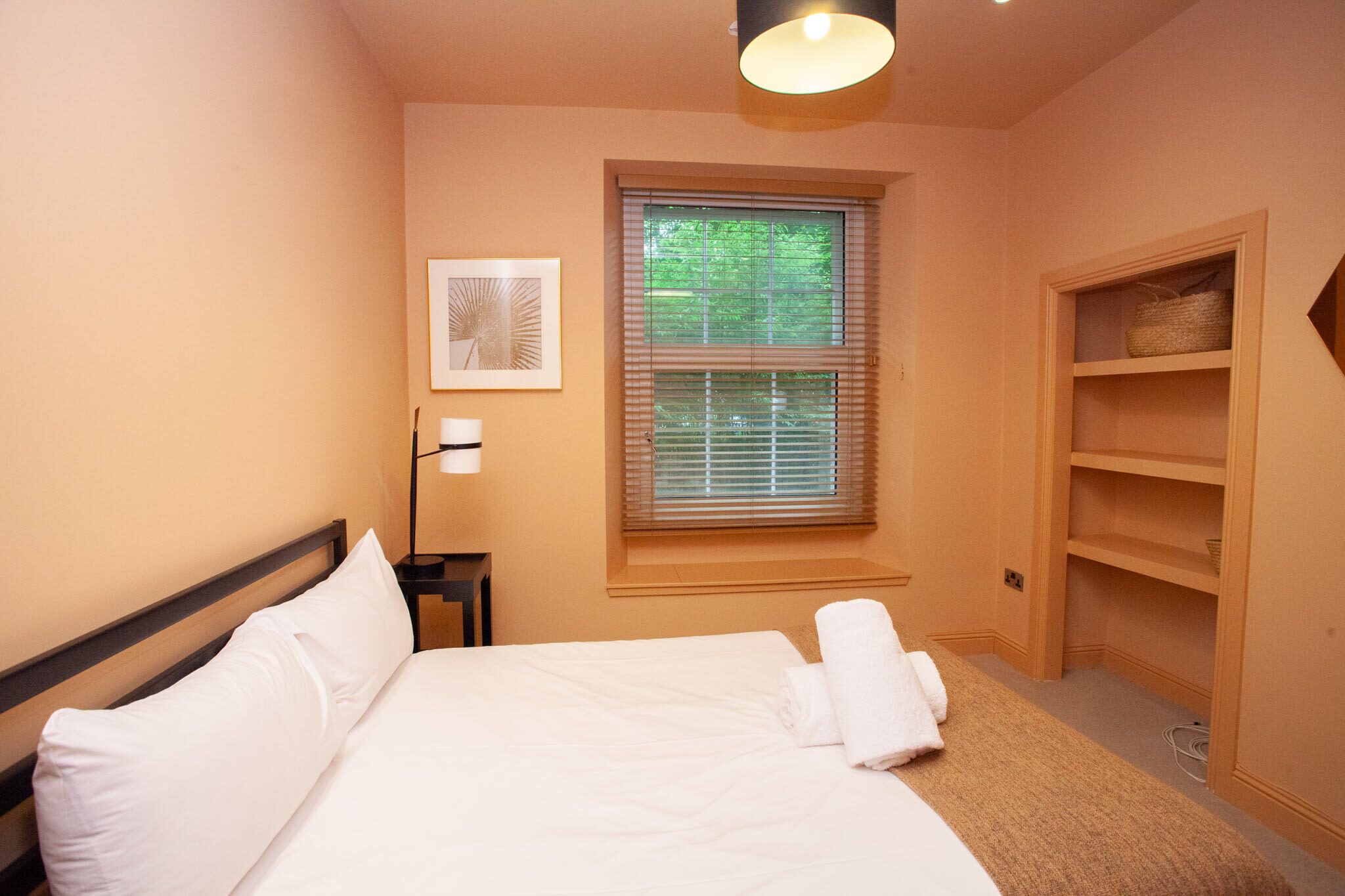 A bedroom with a bed, a bedside table, a lamp, a window with blinds, and a built-in shelf.