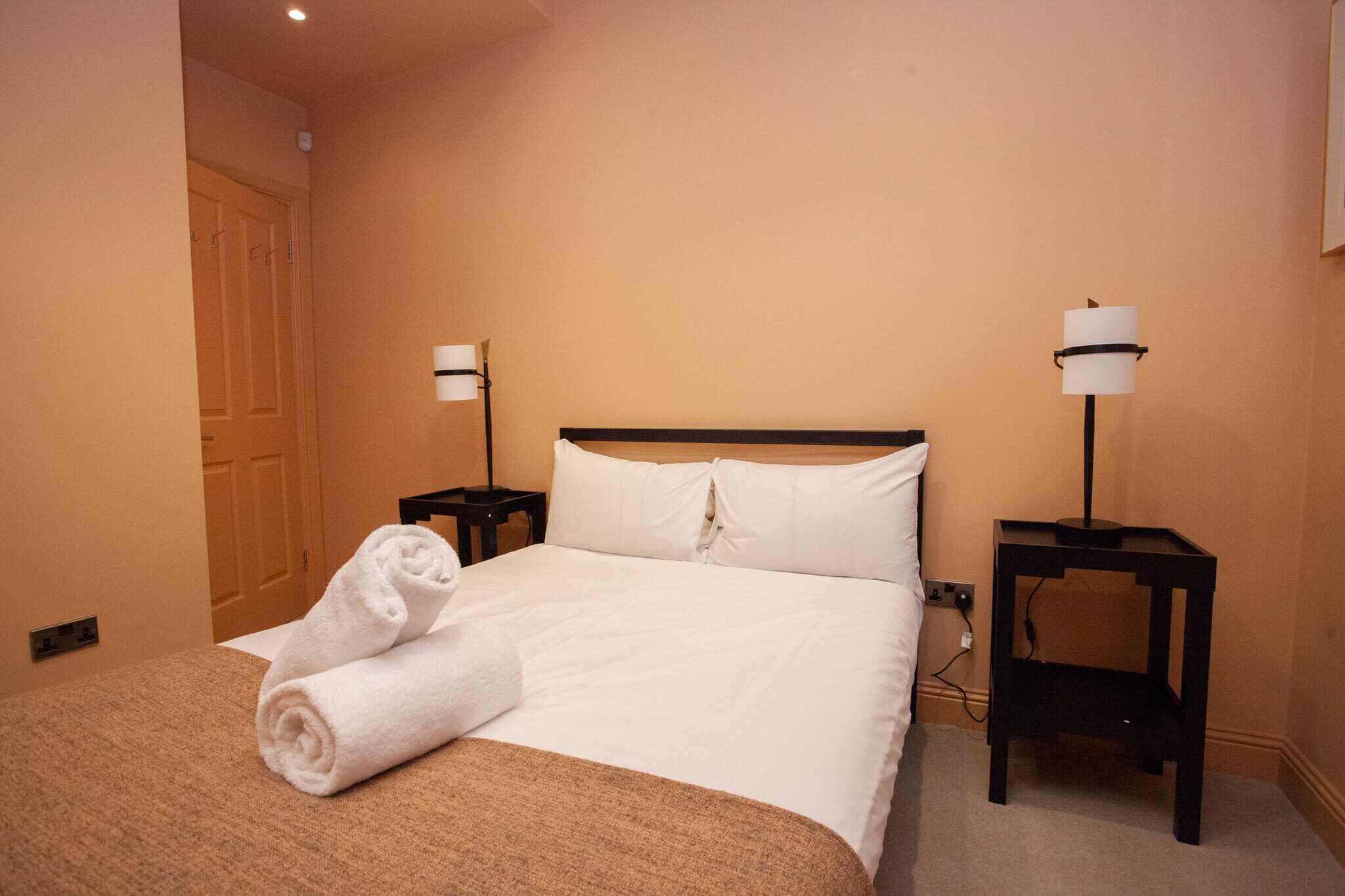 A hotel room with a bed, two bedside tables, a lamp, and a rolled towel on the bed.