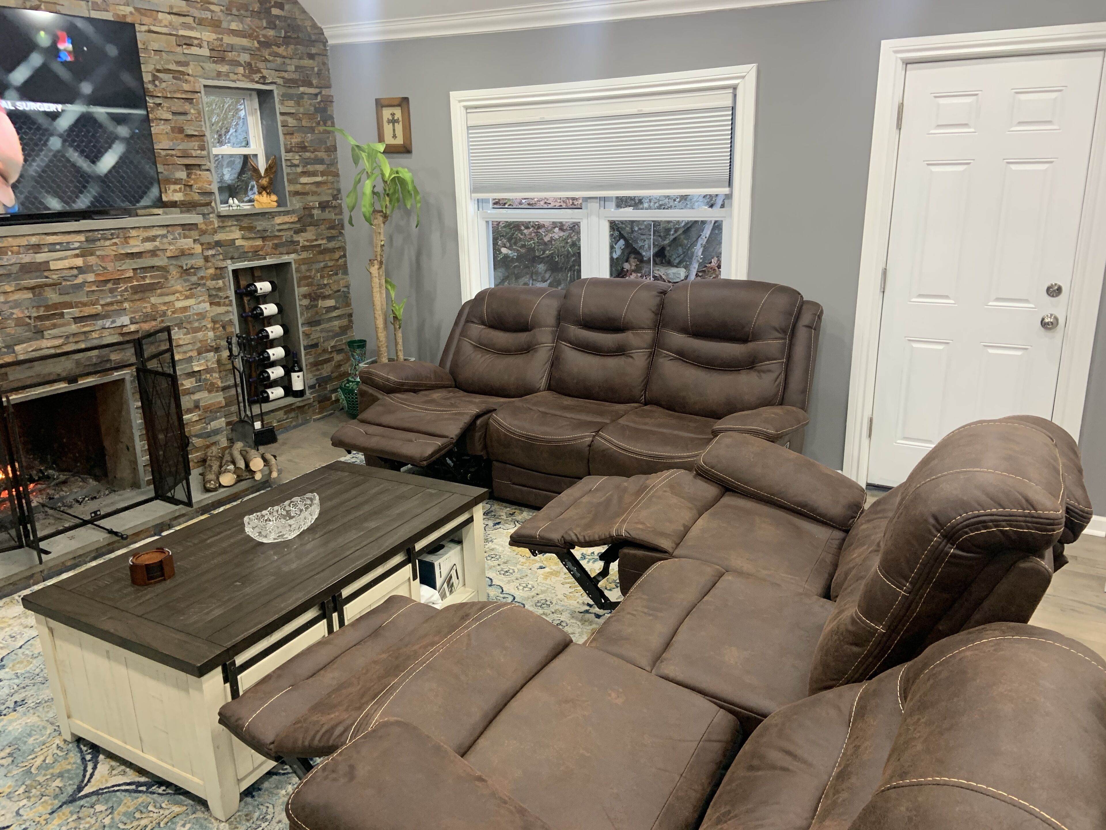 A living room with a stone fireplace, a brown sectional sofa, a coffee table, and a television mounted on the wall.