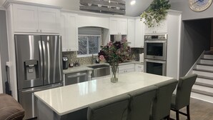 Uma cozinha moderna com armários brancos, eletrodomésticos de aço inoxidável, uma ilha central com banquetas e uma planta decorativa.