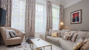 A living room with a beige sofa, a round armchair, a coffee table, and a TV on the left wall. - Charming 3-Bedroom Retreat in Glasgow West End (Glasgow City)