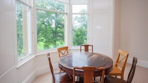 A dining area with a round table and six wooden chairs, large windows with a view of trees, and white paneled walls. - Spacious, 4 Bed Flat Overlooking Park in West End (Glasgow)