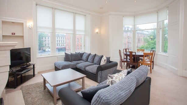 A spacious living room with a sectional sofa, a coffee table, and a TV on a stand. Large windows offer a view of the city. - Spacious, 4 Bed Flat Overlooking Park in West End (Glasgow)