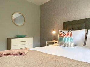 Een netjes opgemaakt bed met patroon kussens, een nachtkastje met een lamp en een dressoir met een kom erop.