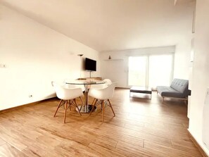 A spacious living area with a round dining table and chairs, a grey sofa, and a flat-screen TV mounted on the wall.