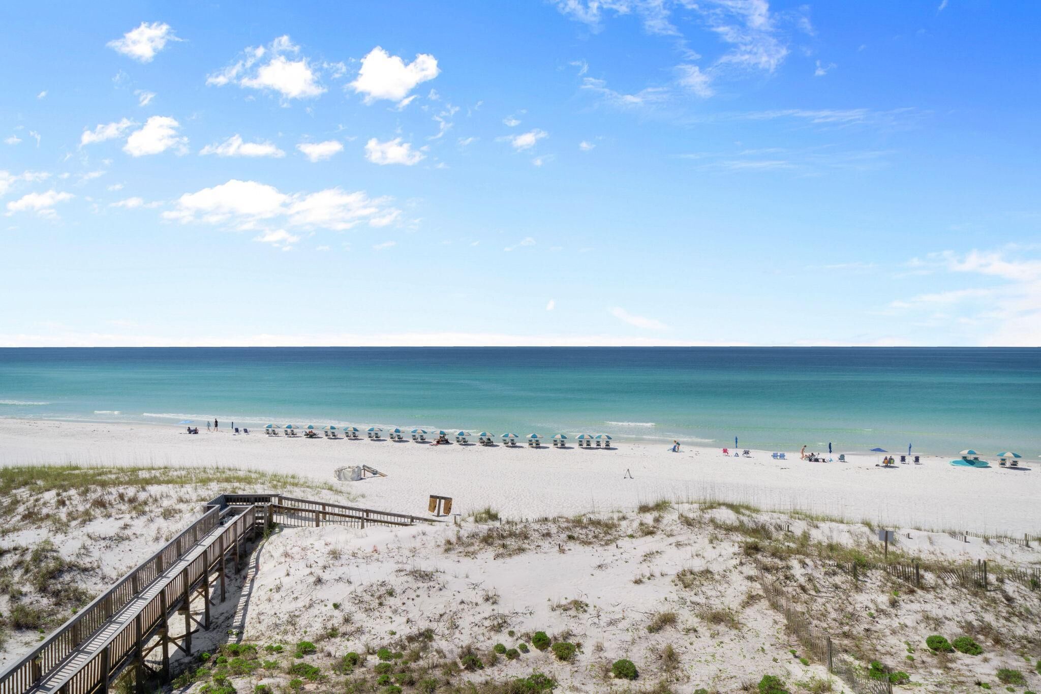 Una spiaggia con una passerella in legno che conduce a un piccolo edificio, dune sabbiose e cielo azzurro terso.