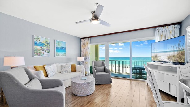 Un salón amplio con sofá, sillones, una mesa de centro y televisor. La habitación tiene ventanas grandes con vistas al océano.