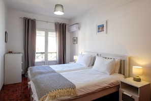 A hotel room with two beds, a bedside table with a lamp, a wardrobe, a window with curtains, and an air conditioning unit.