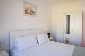 A bedroom with a bed, white pillows, a nightstand with a lamp, a wardrobe, and a framed picture on the wall.