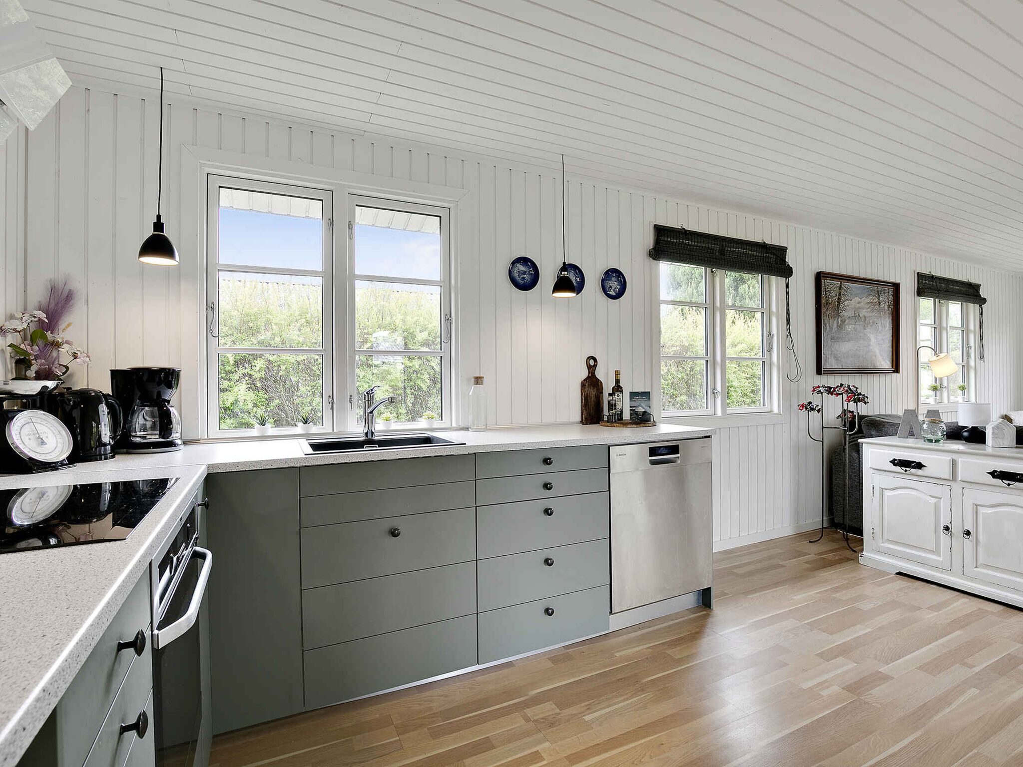 A modern kitchen with white wooden paneling, a large island with grey drawers, a built-in oven, and a window with a view of greenery.