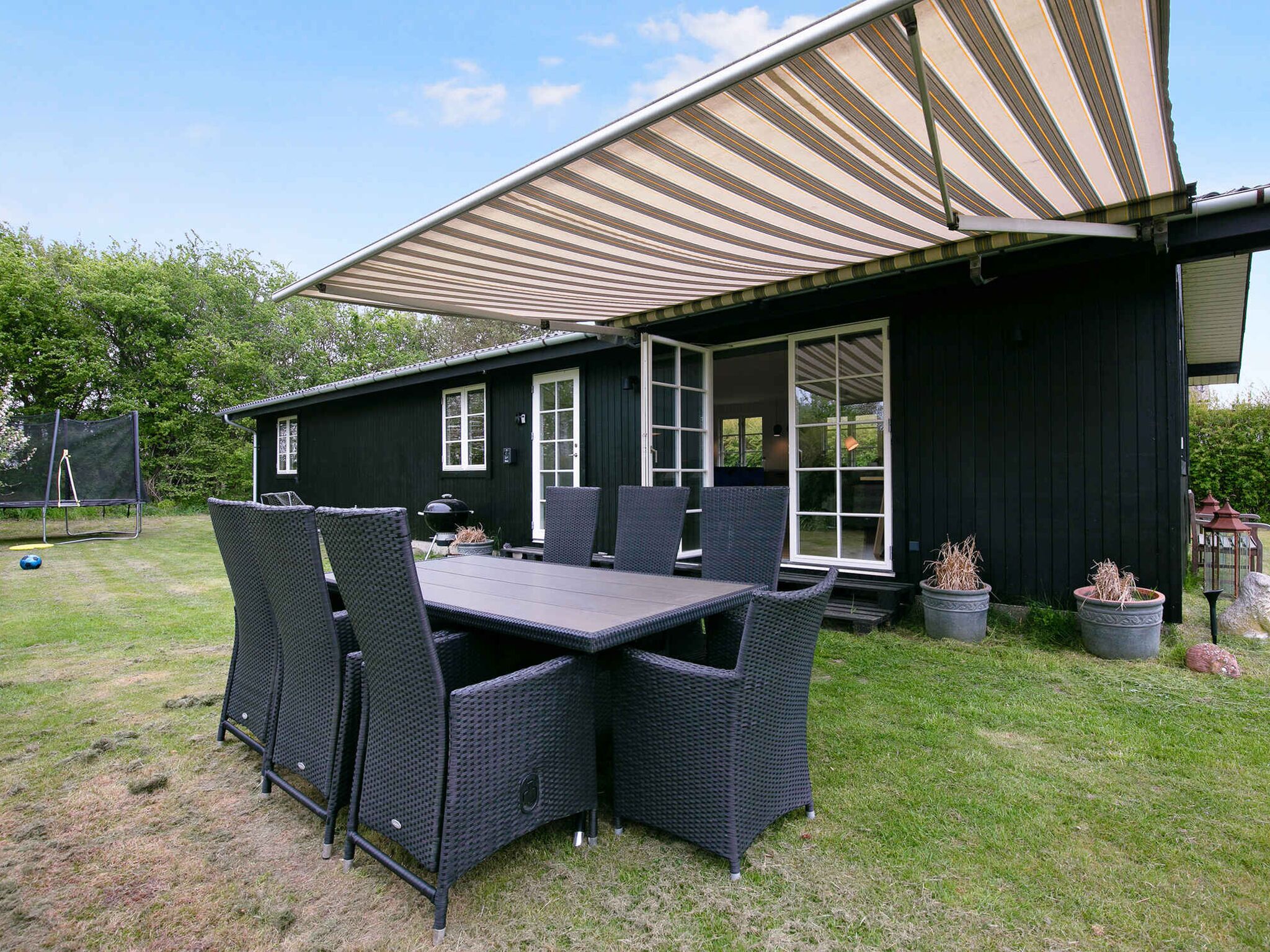 Een zwart huis met een patio, daaraan een tafel, stoelen, een barbecue en potplanten.