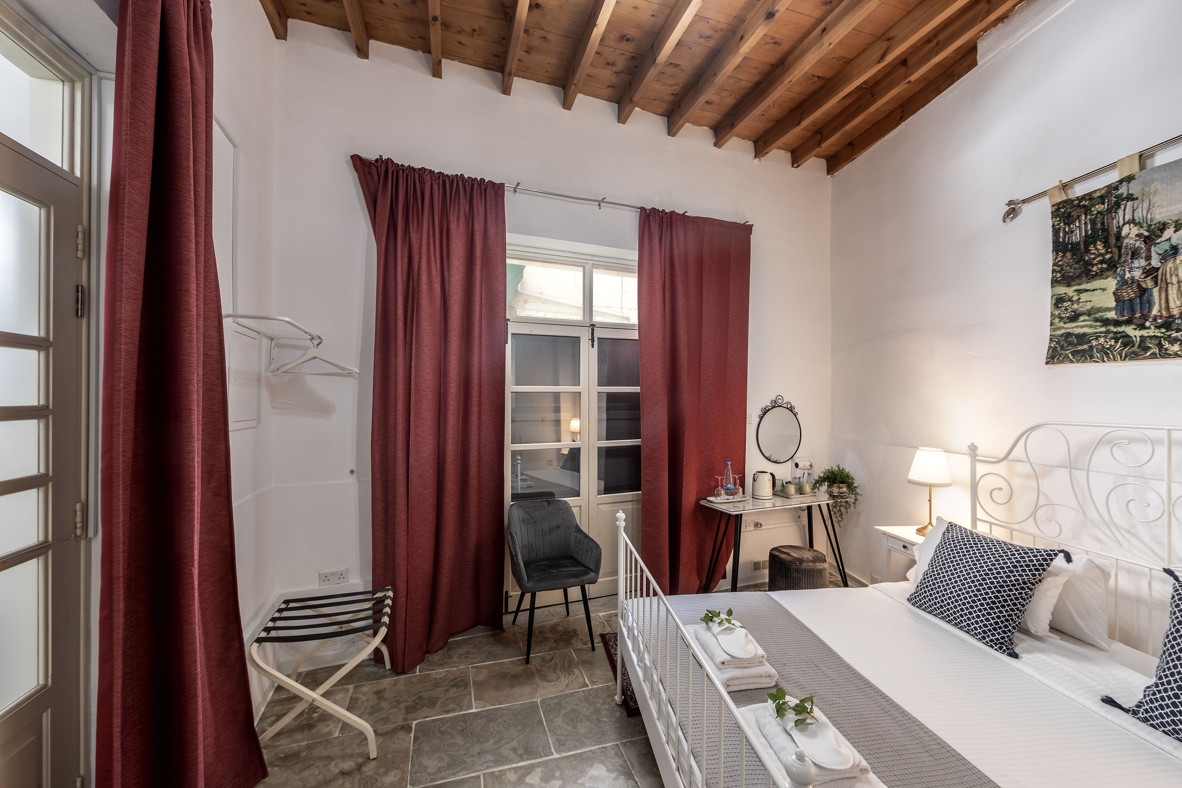 A bedroom with a bed, a chair, a small table, a window with curtains, and a painting on the wall.