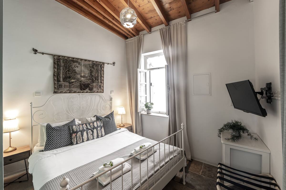 A neatly arranged bedroom with a bed, bedside tables, a mounted TV, and a window with curtains.
