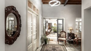 Um hall espaçoso com um espelho grande e ornamentado, vigas de madeira no teto e um lustre moderno. Há áreas de estar confortáveis com cadeiras e uma pequena mesa, e um vaso de planta traz um toque de verde.
