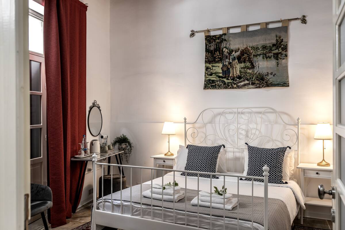 A neatly arranged bedroom with a white metal bed frame, two bedside tables with lamps, a wall-mounted painting, and a red curtain.