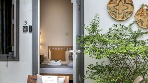 Quarto de hotel com uma cama, mesa de cabeceira e janela. Uma porta dá para o exterior, e uma planta é visível em primeiro plano.