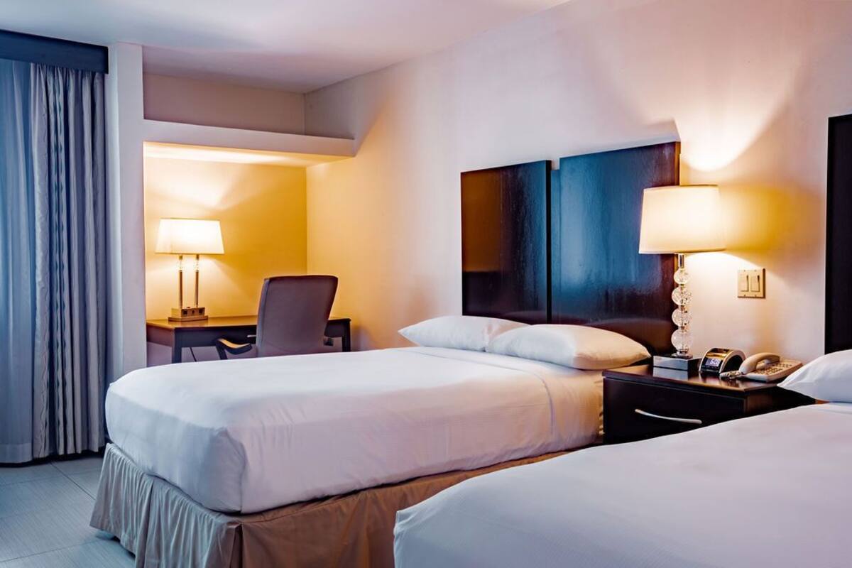 A hotel room with two beds, a desk, and a chair. There are lamps on the desk and bedside tables.