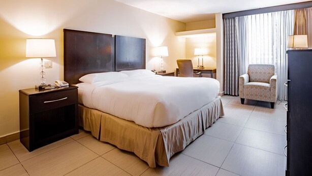 A hotel room with a large bed, bedside tables with lamps, a chair, and a large window with curtains. - Studio Coliving (Panama City)