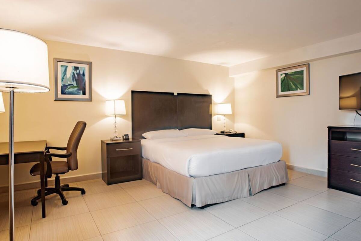 A hotel room with a bed, desk, chair, nightstands, lamps, and artwork on the walls.