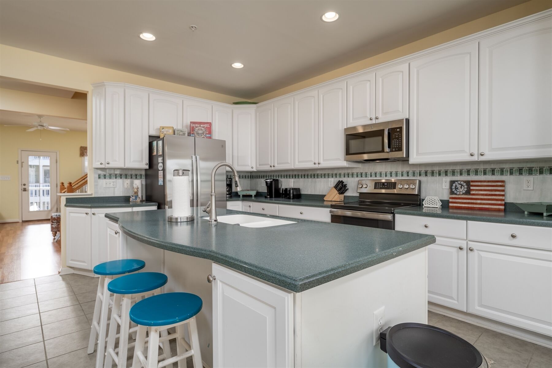 A kitchen with white cabinets, a granite countertop, a stainless steel refrigerator, a microwave, and a coffee maker. There are three blue bar stools.