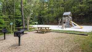 Une aire de jeux avec des balançoires, un toboggan et une table de pique-nique, située dans un espace boisé.