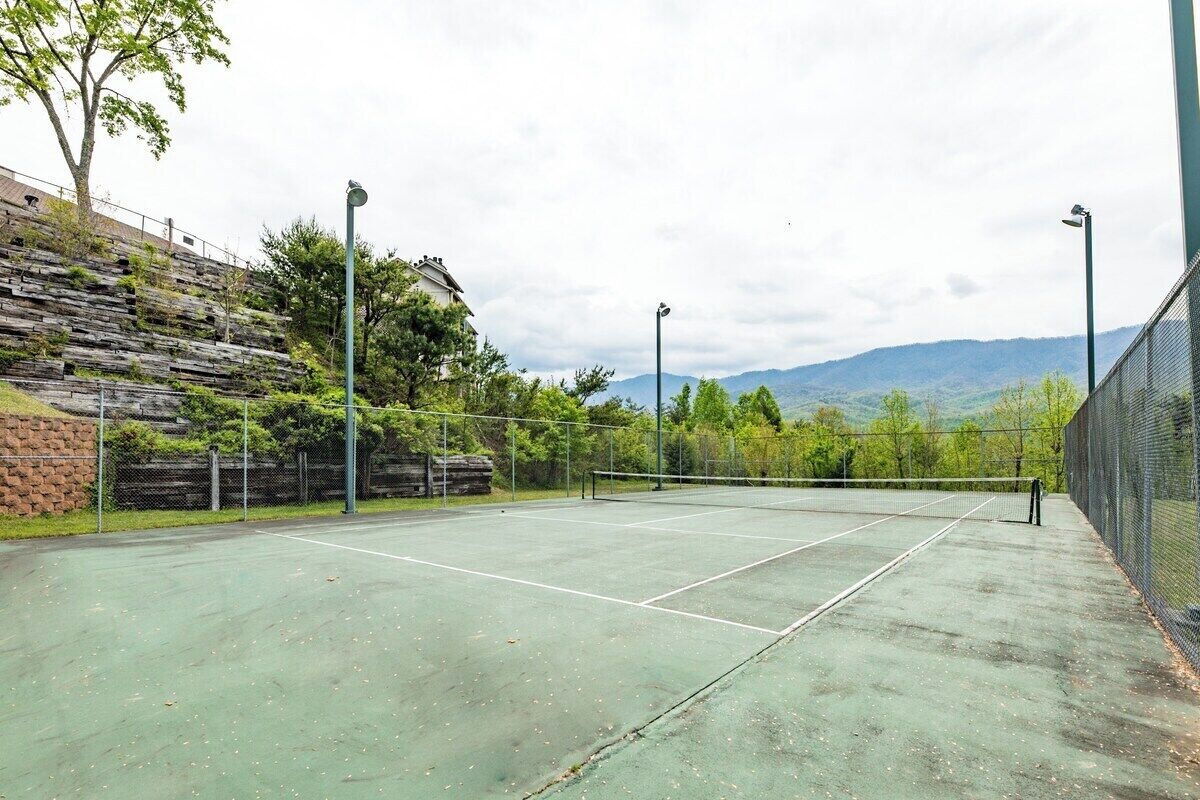 An outdoor tennis court with a net, surrounded by a fence, and a hillside in the background.