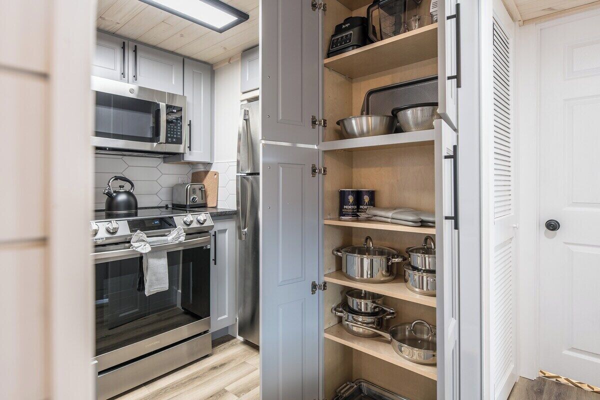 A kitchen with stainless steel appliances, a microwave, a kettle, and a built-in oven. There is a pantry with shelves holding pots and pans.