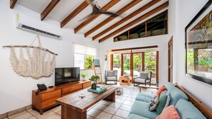 Un salón amplio con techo de madera, televisor, mesa de centro de madera, sofá y un ventanal con vistas al verdor.