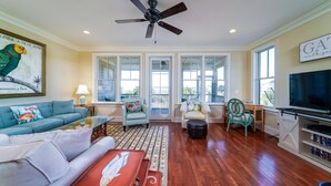 A spacious living room with a ceiling fan, large windows, a television, and a bookshelf. - Stunning Gem! Amazing Marsh/Tidal Views. Amenity access included! (Seabrook Island)