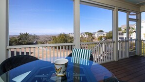 A covered outdoor area with a table and chairs, a cup on the table, and a view of the beach and houses in the background. - Stunning Gem! Amazing Marsh/Tidal Views. Amenity access included! (Seabrook Island)